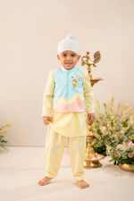 A young child wearing a traditional Indian outfit consisting of a light yellow kurta and matching pants, a light blue embroidered vest, and a white beanie hat, standing in a studio setting with decorative props.