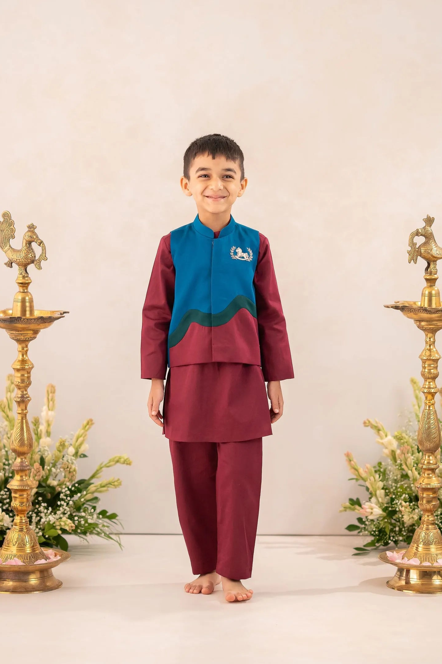 A young boy stands barefoot in the center of a studio setup, wearing a traditional Thai outfit consisting of a maroon kurta and matching trousers with a teal and green embroidered yoke. He is flanked by two ornate gold oil lamps and floral arrangements against a warm beige background.