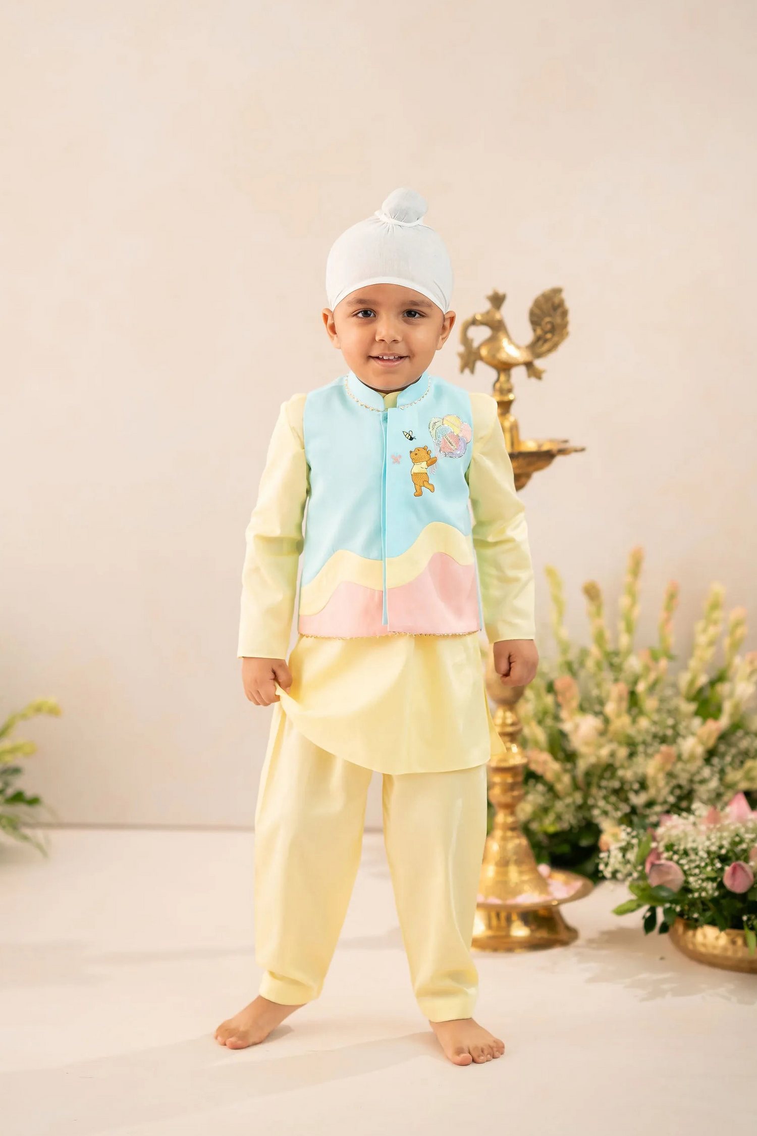 A young child wearing a traditional Indian outfit consisting of a light yellow kurta and matching pants, a light blue embroidered vest, and a white beanie hat, standing in a studio setting with decorative props.