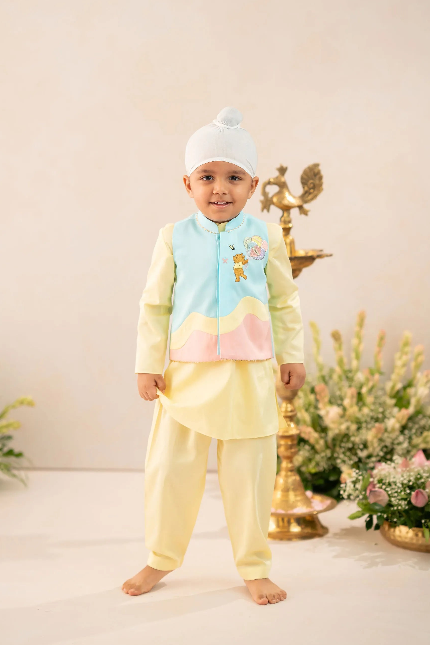 A young child wearing a traditional Indian outfit consisting of a light yellow kurta and matching pants, a light blue embroidered vest, and a white beanie hat, standing in a studio setting with decorative props.